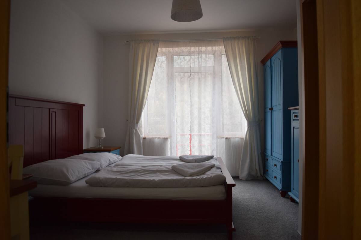 A bedroom with a bed, a nightstand, a window with curtains, and a blue wardrobe.