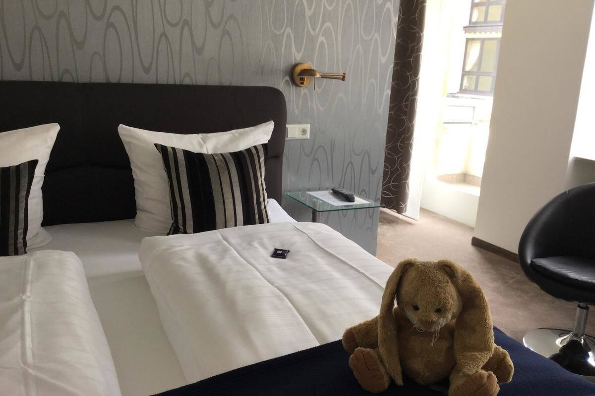 A hotel room with a bed, a teddy bear, and a chair.
