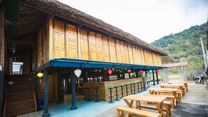 A wooden building with a thatched roof, outdoor seating area with wooden tables and benches, and a bar counter with stools. - Ngoc Son homestay (Ha Giang 1)