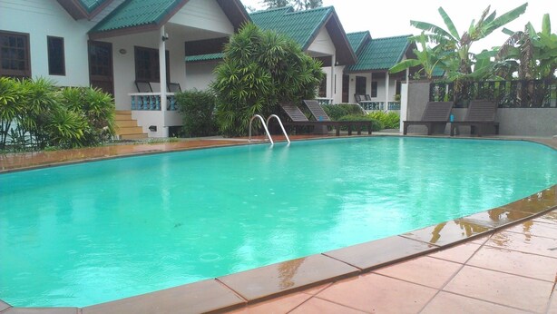 A swimming pool with a tiled edge, surrounded by a building with a green roof and a covered patio area. - Surin House (Choeng Thale)