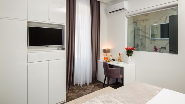 A hotel room with a bed, a desk with a chair, a TV, a window with a view, and an air conditioning unit. - Dream Luxury Rooms (Split)