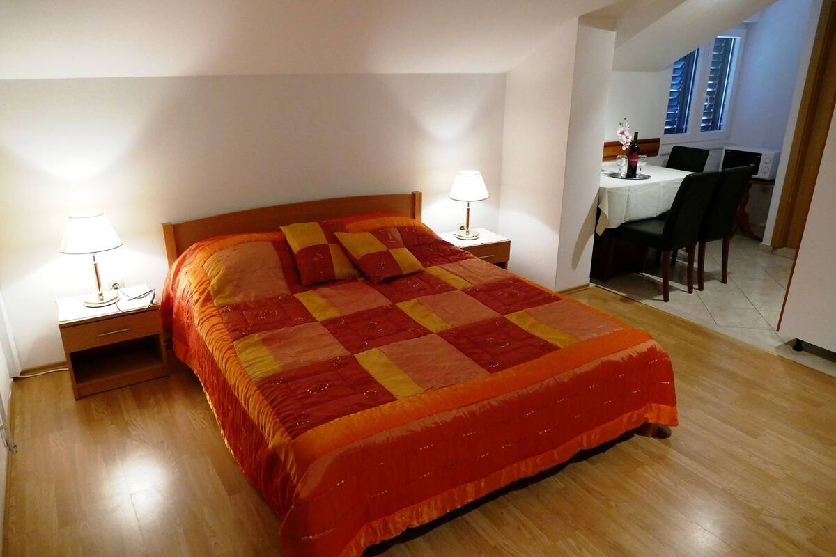 Una cama bien tendida con un colcha rojo y amarilla, dos mesitas de noche con lámparas y una pequeña zona de comedor con una mesa y sillas.