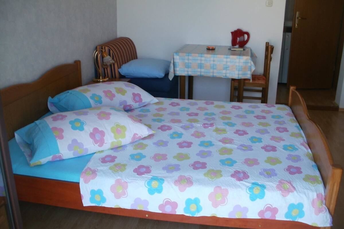 A bed with a floral bedspread, a bedside table with a red kettle, and a chair.