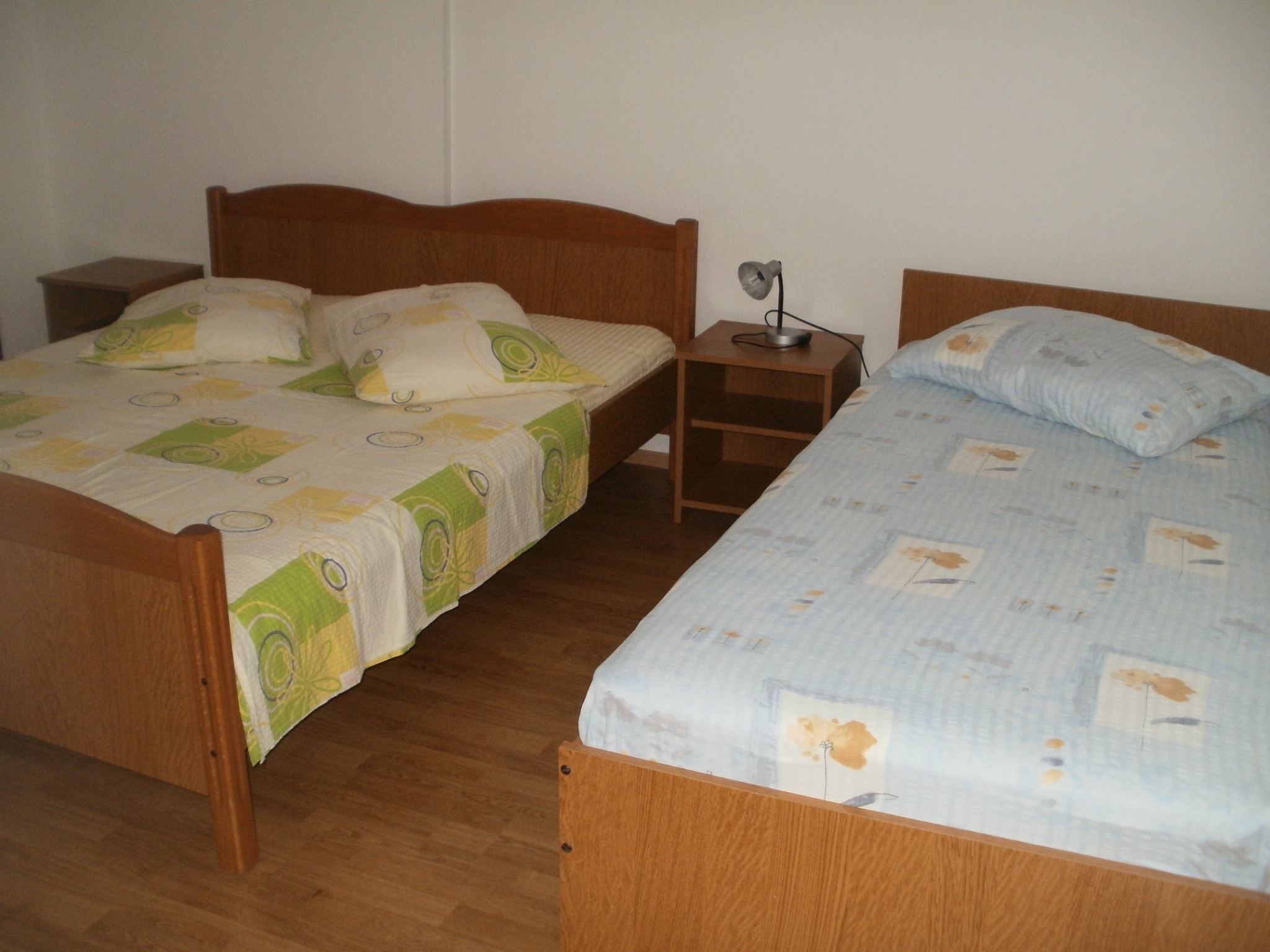 A room with two wooden beds, each with a different patterned bedspread. There is a nightstand with a lamp between the beds.