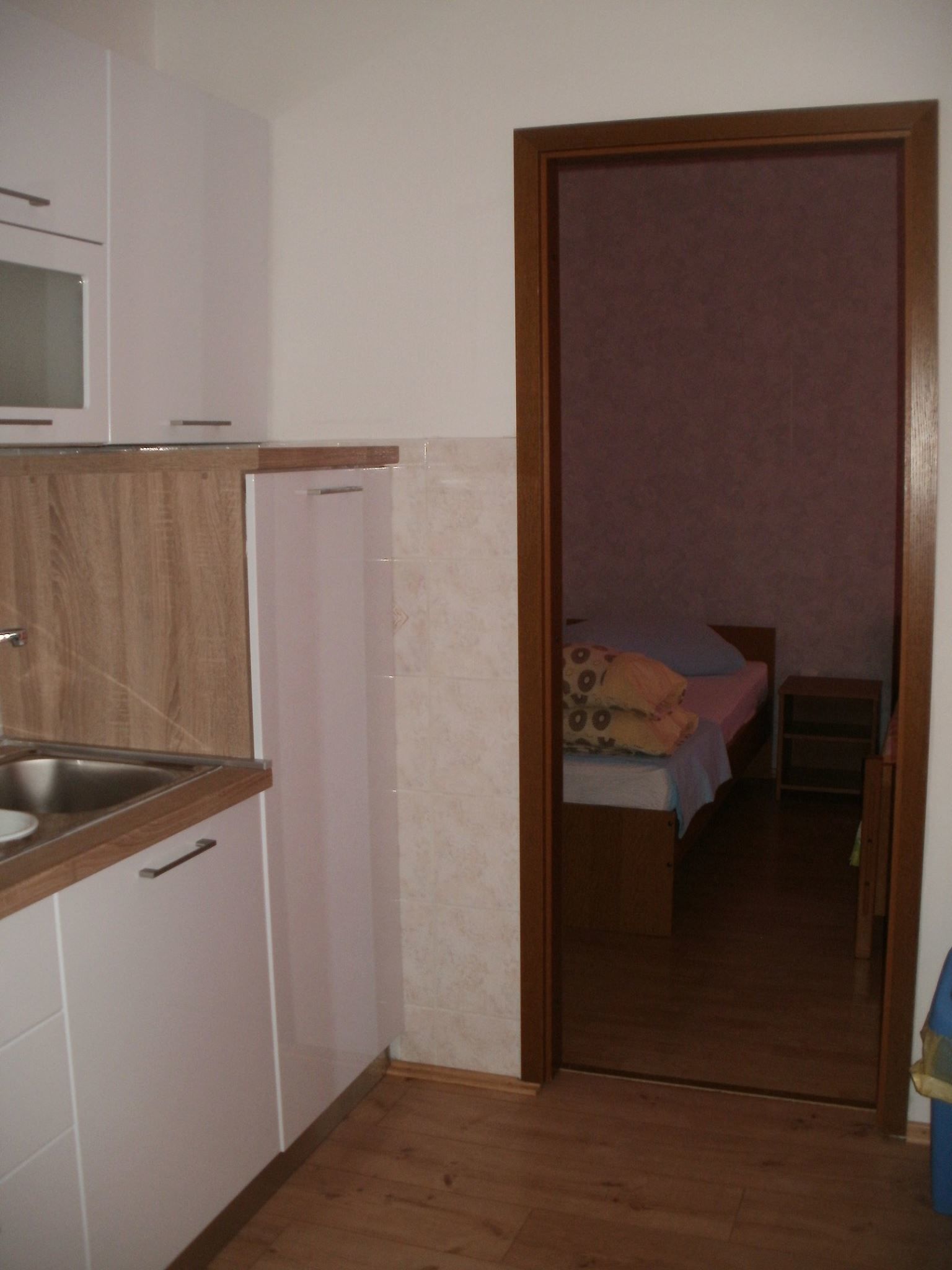 Cocina compacta con microondas, pileta y muebles de madera. Se ve un dormitorio con cama y mesita de noche a través de una puerta abierta.