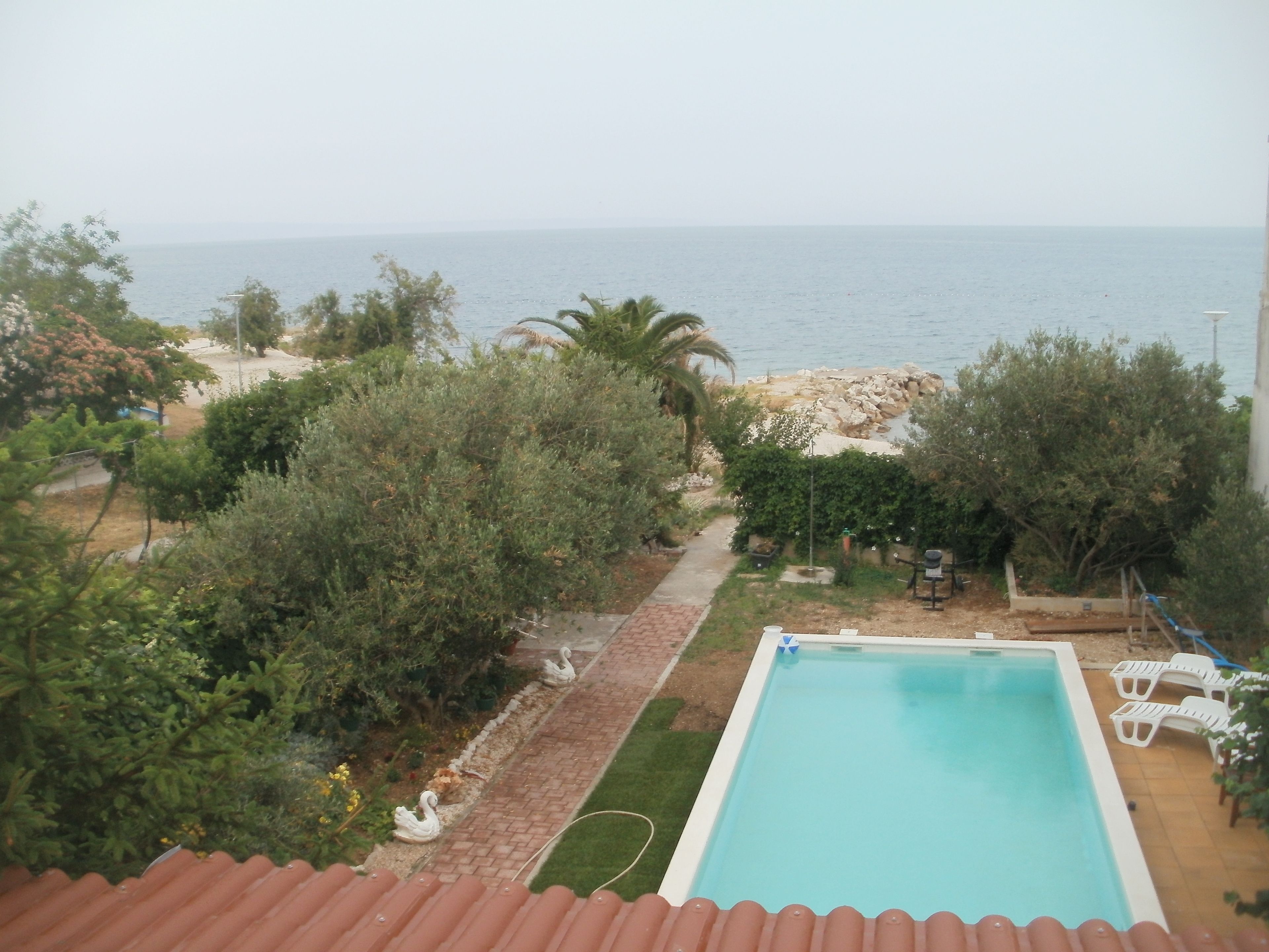 A swimming pool with a view of the sea, surrounded by greenery and a patio area with outdoor furniture.