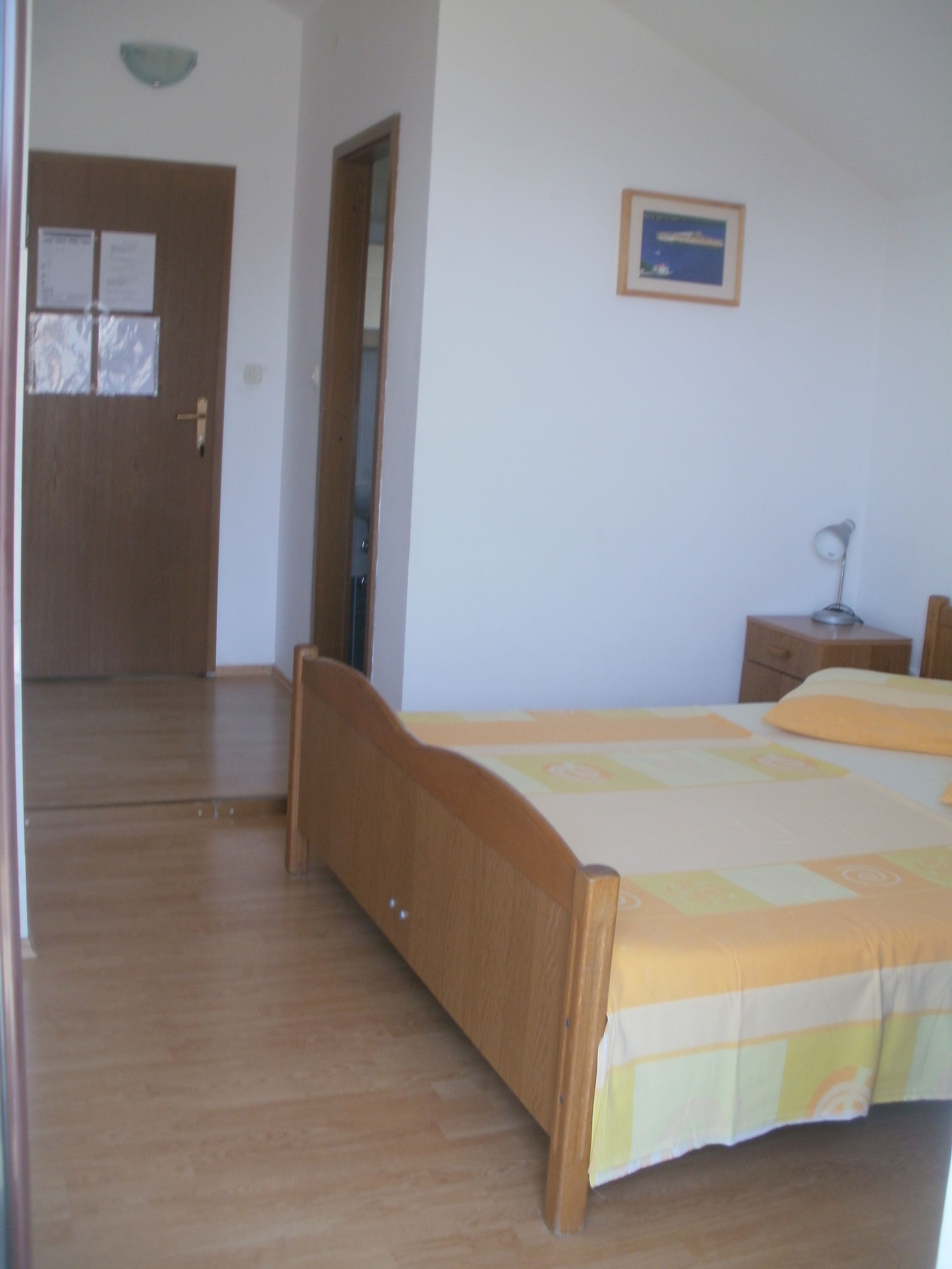 A bedroom with a wooden bed, a nightstand, a desk, and a chair. There is a door with a mirror and a window with blinds.