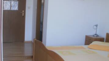 A bedroom with a wooden bed, a nightstand, a desk, and a chair. There is a door with a mirror and a window with blinds.