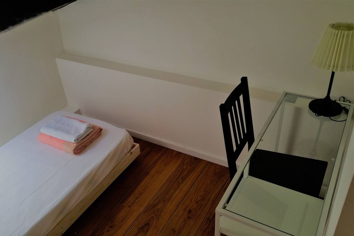 Ein kleines Zimmer mit einem Einzelbett, Holzboden, Schreibtisch mit Lampe und einem Stuhl.