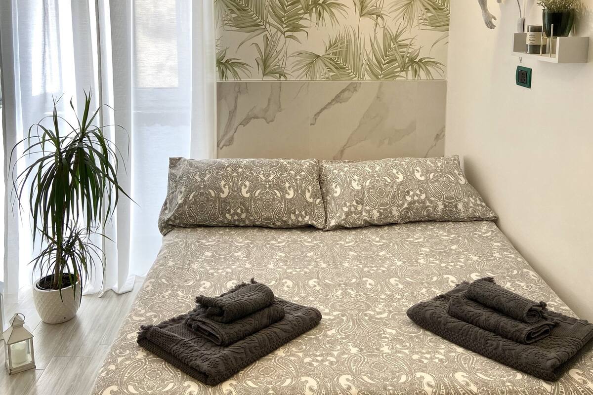 Quarto com cama, roupa de cama estampada, planta em vaso, luminária fixada na parede e objeto decorativo na parede.