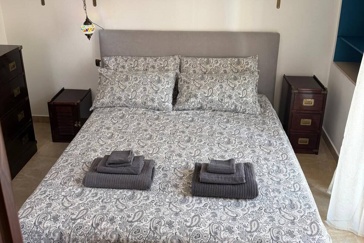 Quarto com cama, mesas de cabeceira, luminária pendente e prateleira na parede.