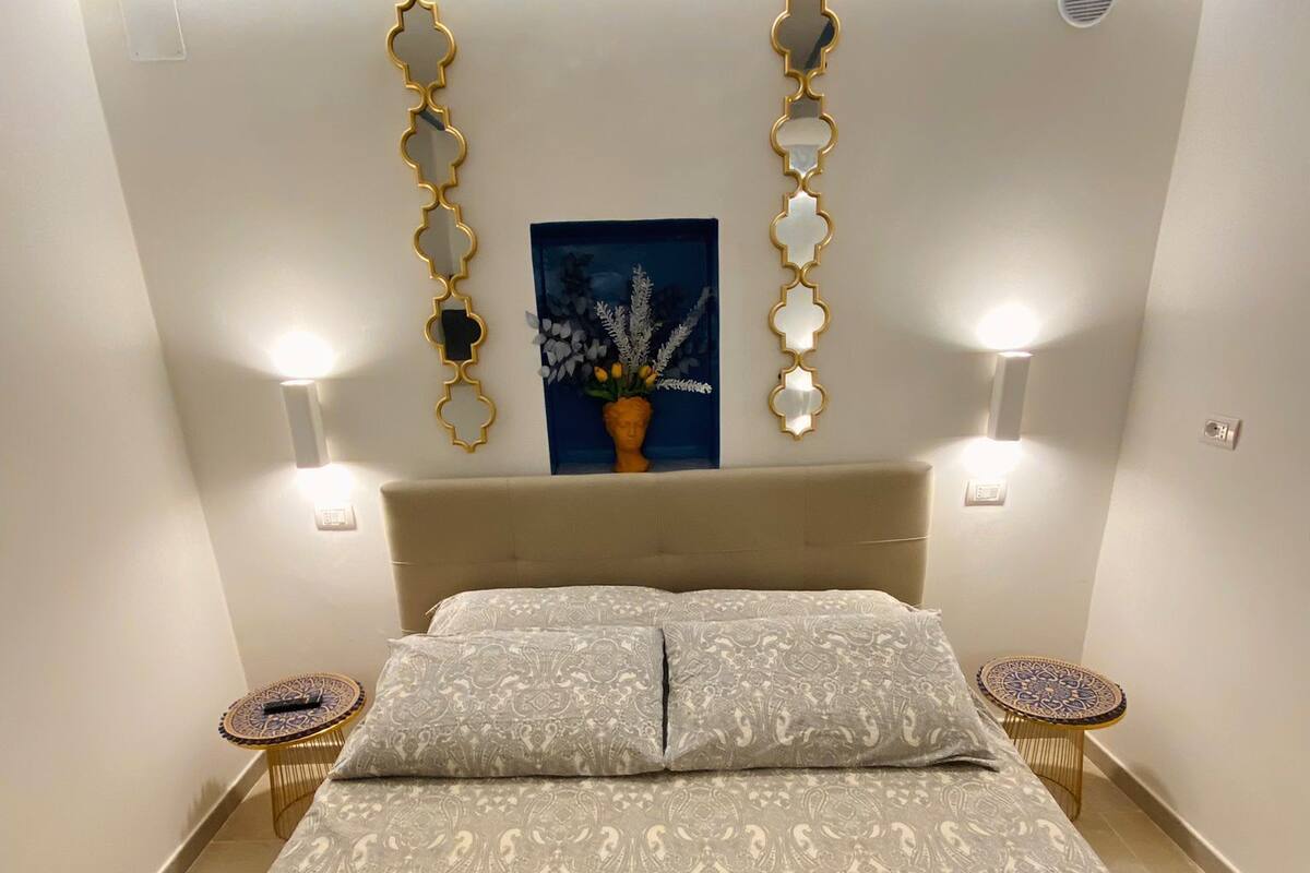 Quarto com uma cama grande, duas mesas de cabeceira, arandelas e uma composição decorativa de espelhos.