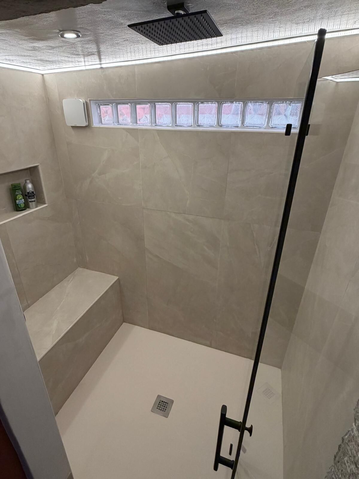 Una ducha con puerta de cristal, un banco y un estante con artículos de aseo.