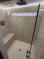 Una ducha con puerta de cristal, un banco y un estante con artículos de aseo.