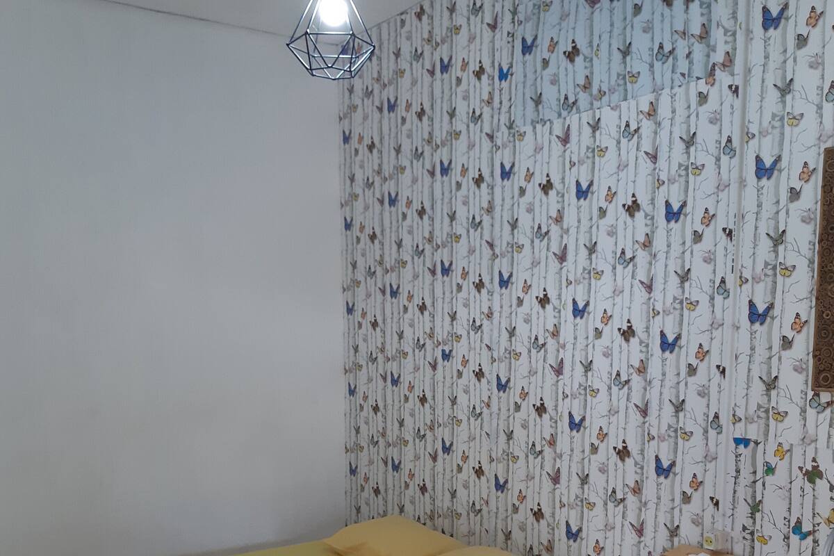 A room with a bed, a hanging light fixture, a patterned wallpaper, and a small shelf with items.