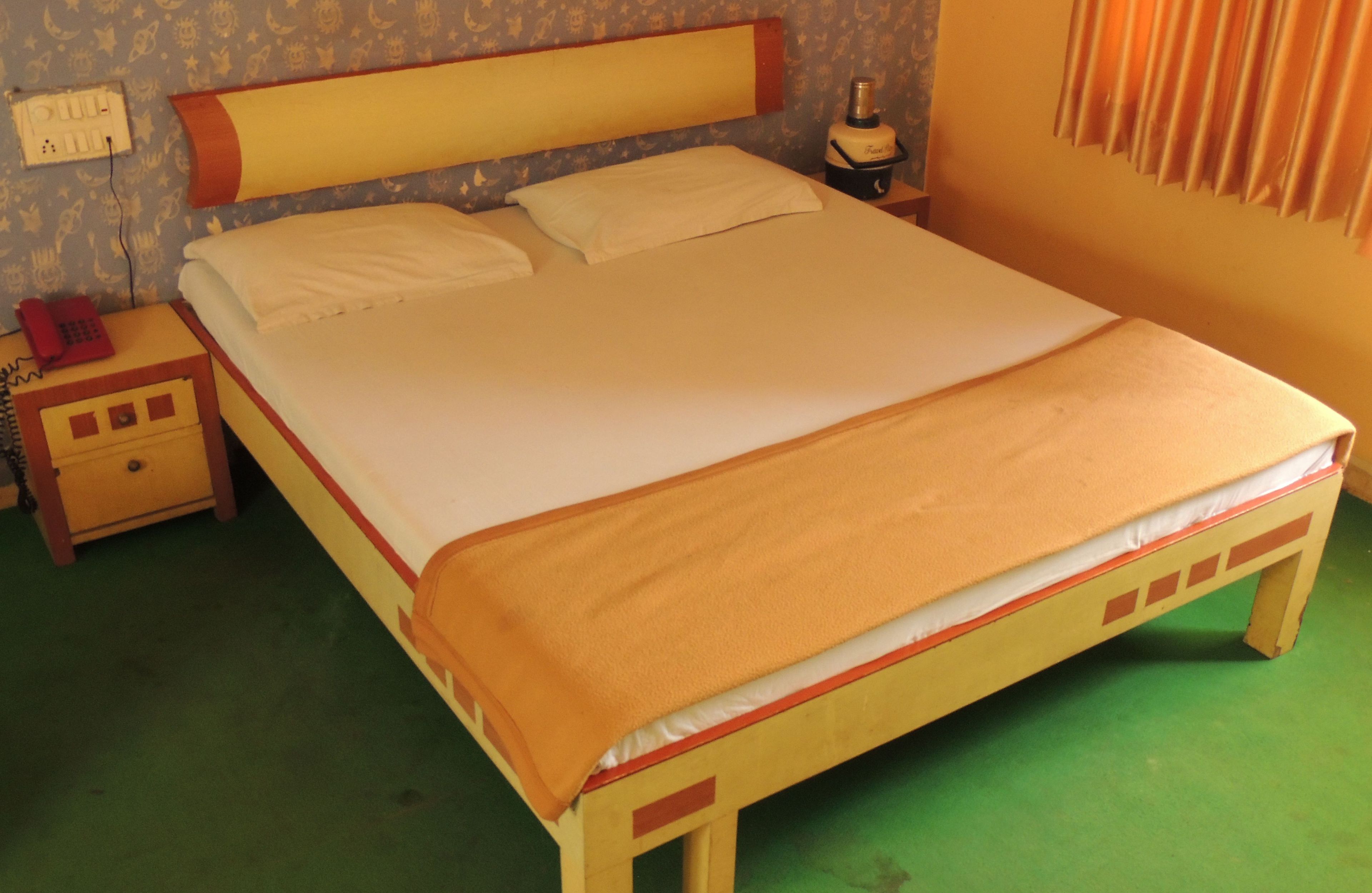 Un lit simple avec un cadre en bois et un matelas beige. Il y a une table de chevet avec un téléphone rouge et une lampe à côté du lit. La pièce est dotée d’une moquette verte et d’un papier peint à motifs.