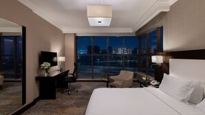Een hotelkamer met een groot bed, een bureau met stoel, een televisie en uitzicht op een stadsbeeld 's nachts.