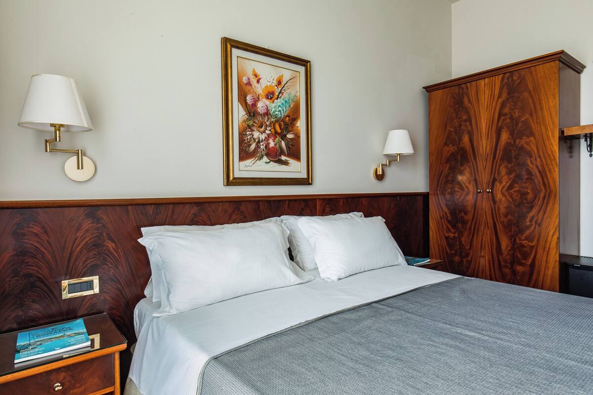 A neatly made bed with white linens, a wooden headboard, and a framed floral artwork above it. There are two bedside lamps and a nightstand with a book and a remote control.