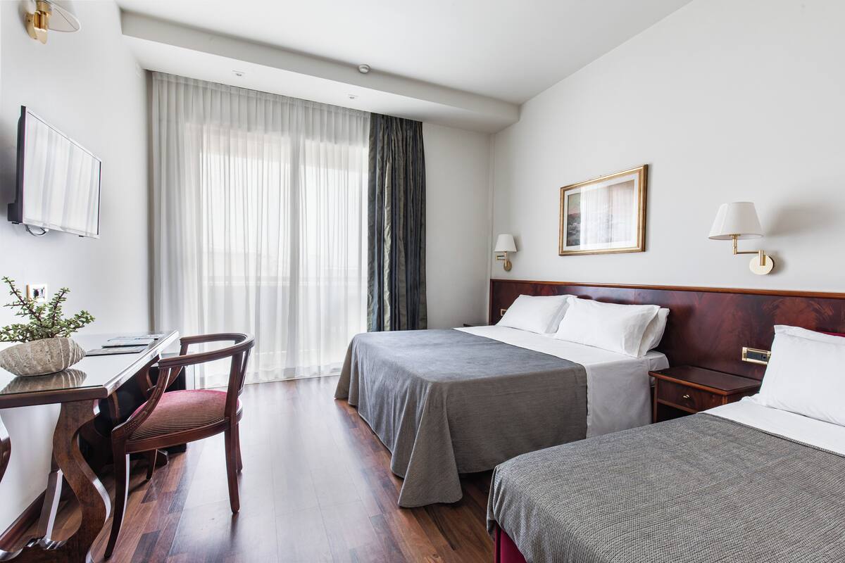 A hotel room with two beds, a desk with a chair, a TV, and a window with curtains.