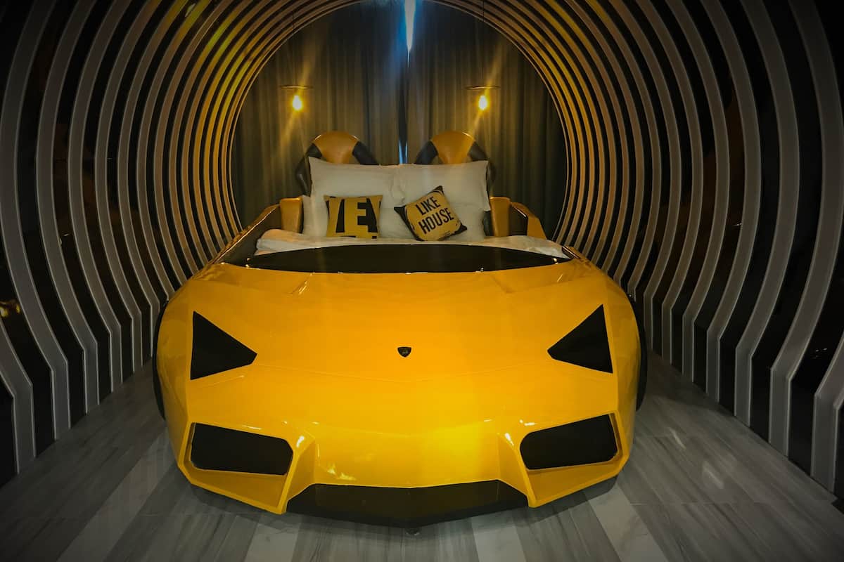 A bed with a unique design resembling a yellow sports car, set in a room with a tunnel-like architectural feature and a modern, striped wall design.