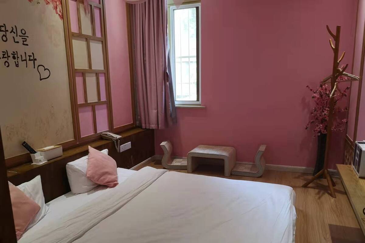A hotel room with a bed, a bedside table, a mirror, a window with curtains, a small stool, and a decorative plant.
