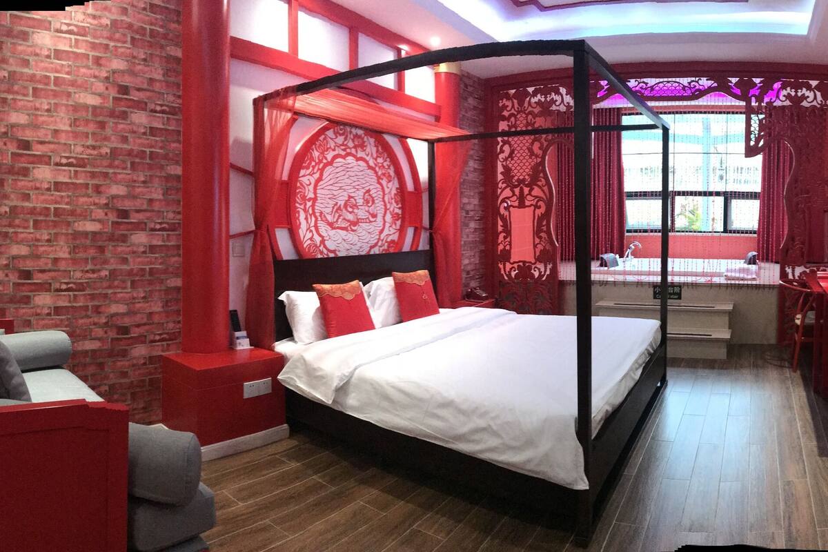 A modern bedroom with a four-poster bed, a red accent wall, and a brick wall.