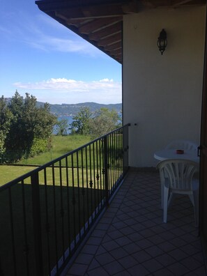 A balcony with a view of a body of water and hills, a white plastic chair, and a small table with a red object on it. - Garni Marika (Gargnano)