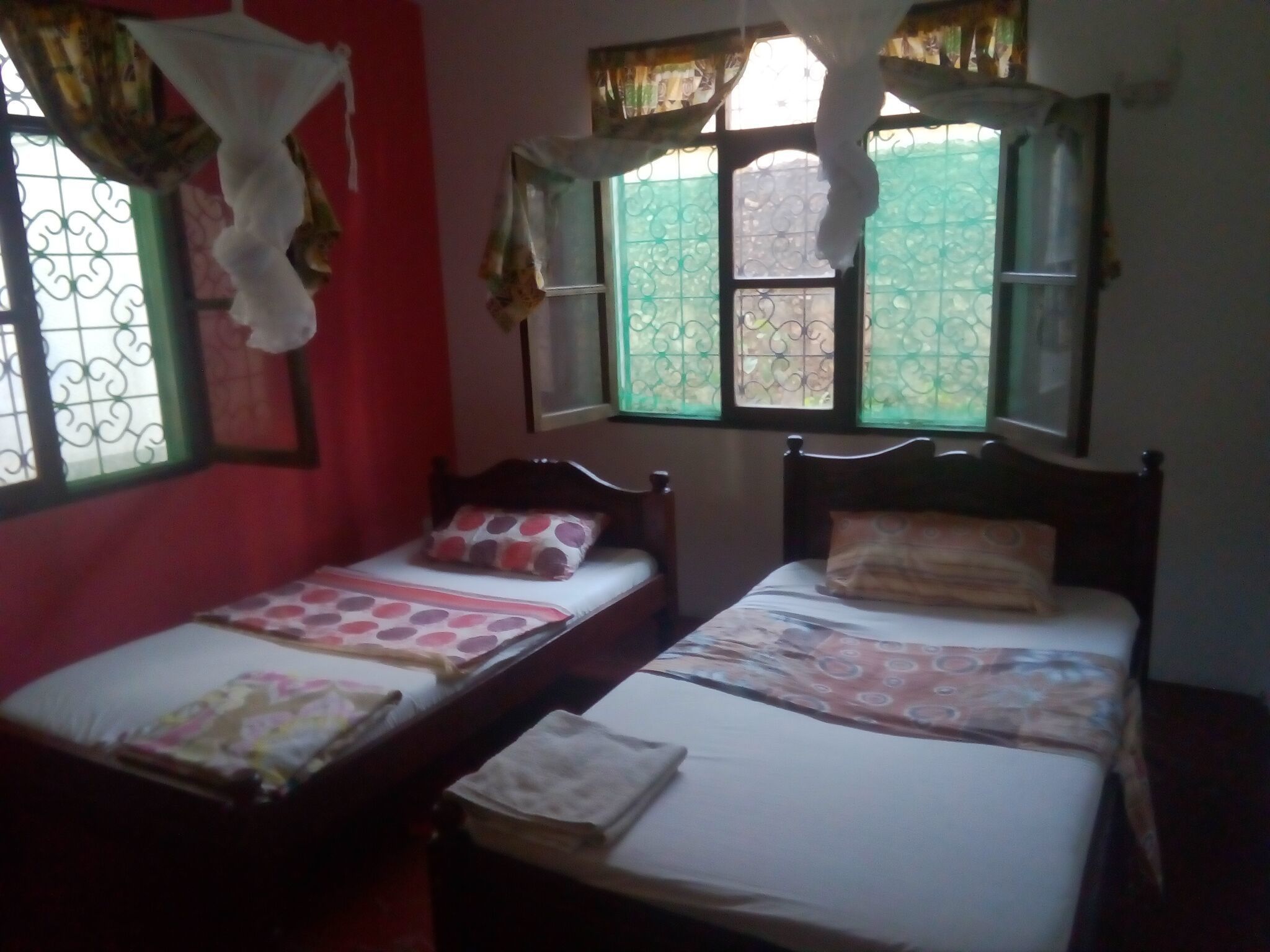 A room with two single beds, each with a pillow and a blanket. There are windows with green curtains and a decorative pattern.