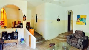 A spacious living area with a sofa, a glass coffee table, and a seating area with a patterned cushion. There is a staircase leading to another level, and a painting on the wall. - Akogo House (Mombasa)