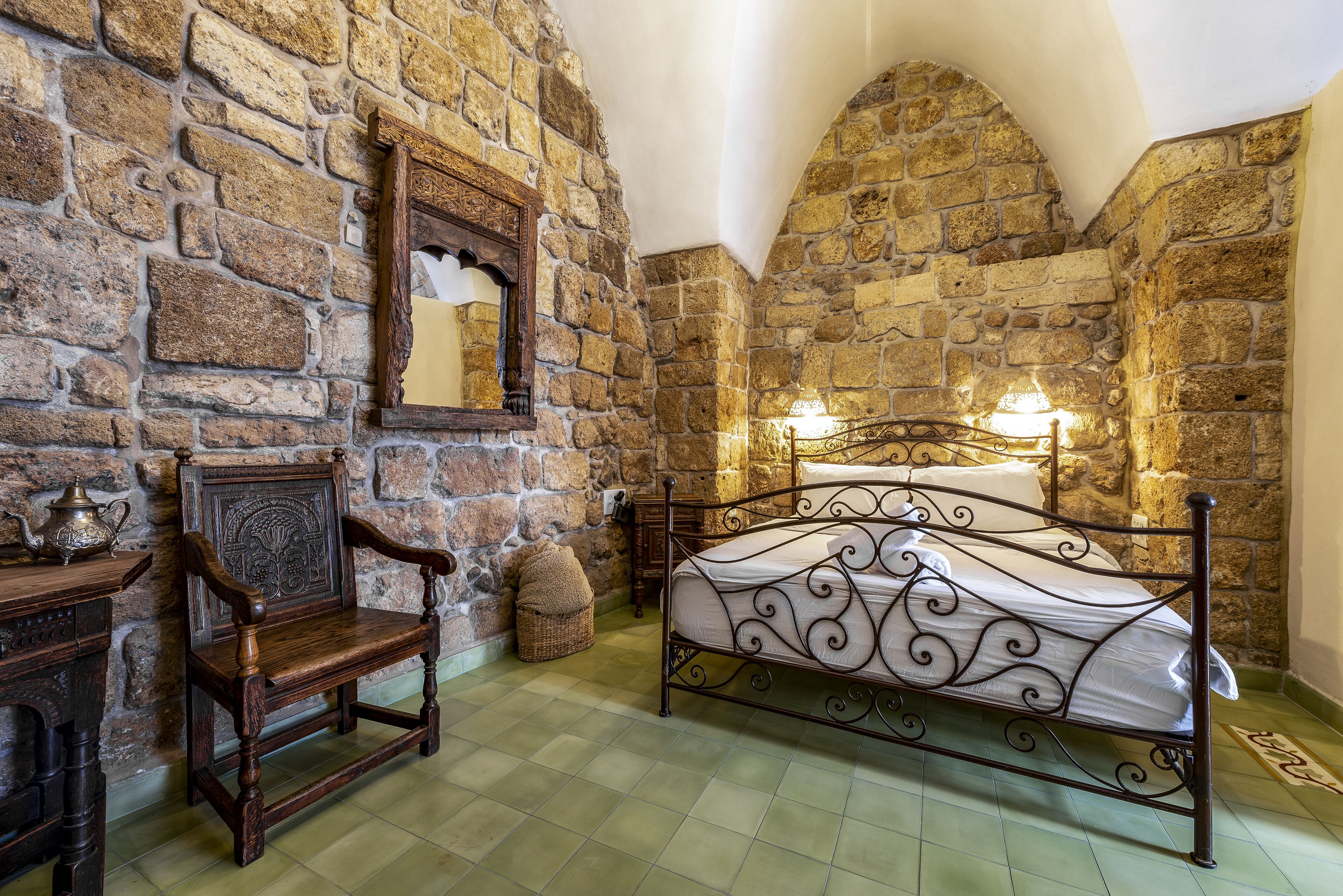 A stone-walled bedroom with a metal bed frame, a wooden chair, and a small table with a lamp.