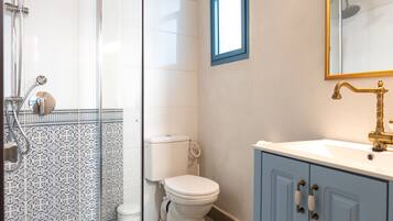 Baño con ducha de vidrio, inodoro, lavamanos con grifo dorado y espejo con marco dorado.