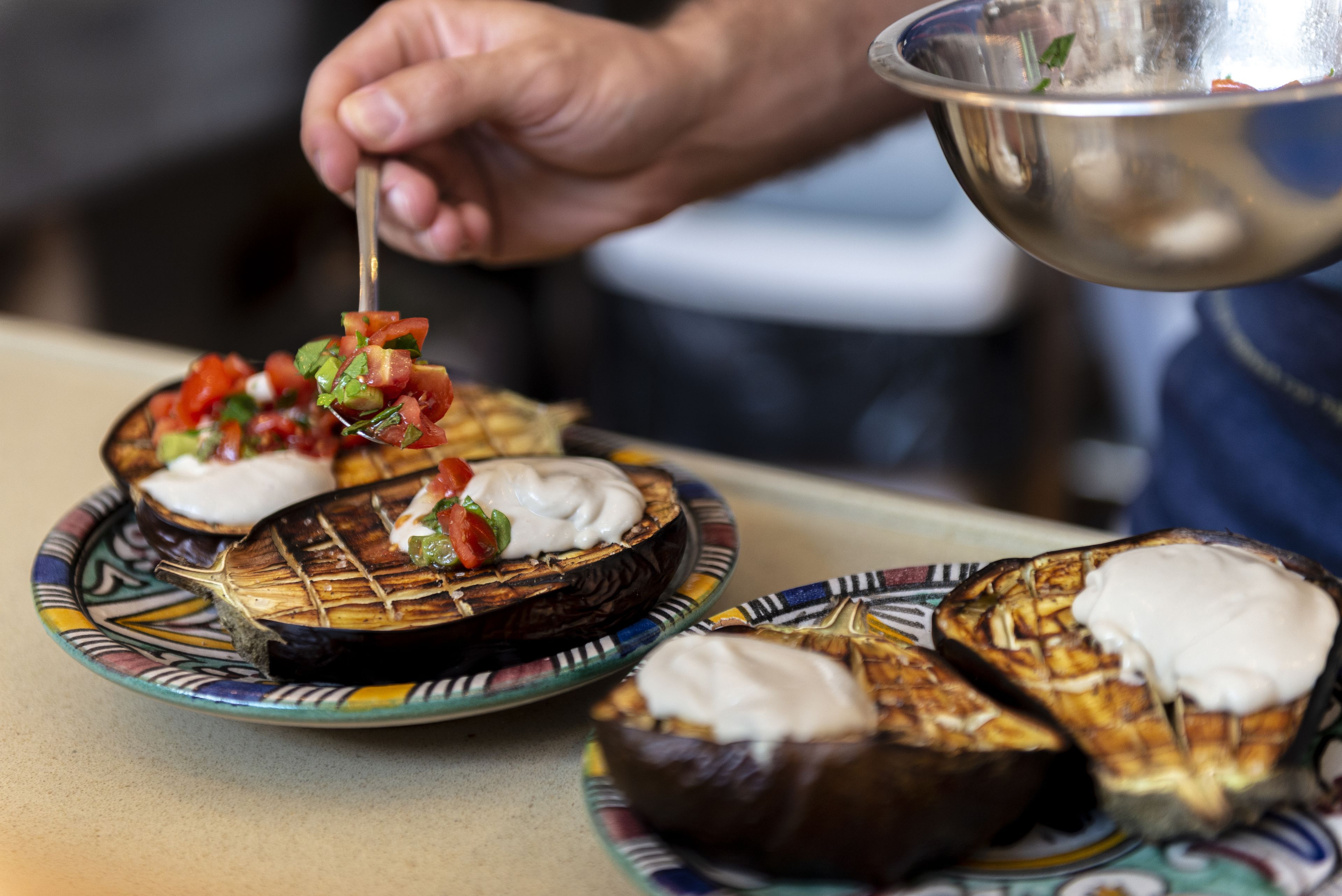 Une personne prépare un plat composé de demi-aubergines grillées, nappées d’une sauce crémeuse et d’un mélange de tomates fraîches.