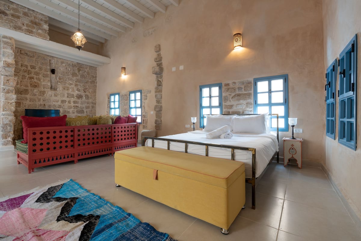 A bedroom with a stone wall, a bed with white linens, a red sofa, a yellow chest of drawers, and a blue window.