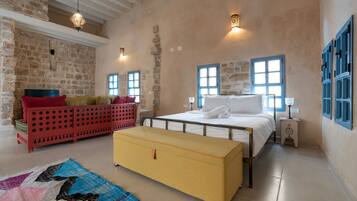A bedroom with a stone wall, a bed with white linens, a red sofa, a yellow chest of drawers, and a blue window.