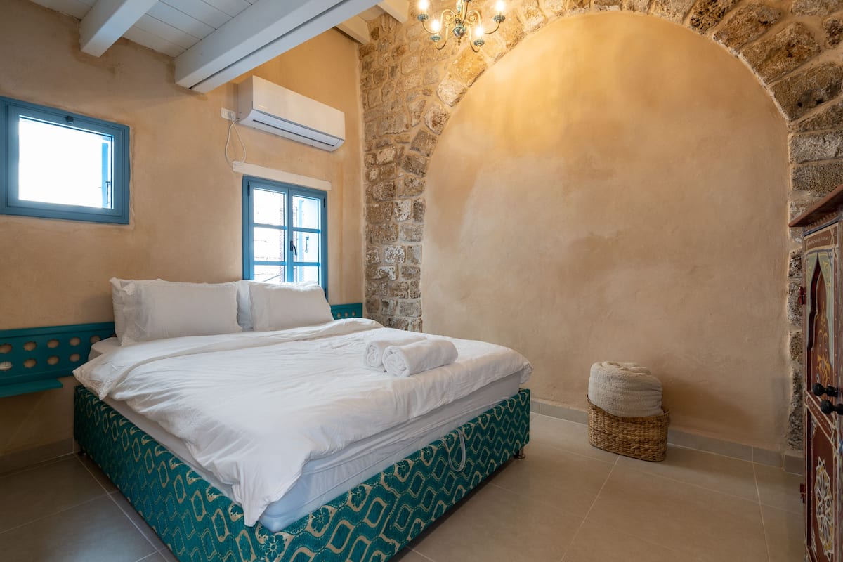 A bedroom with a bed, a window, an air conditioning unit, and a stone wall.