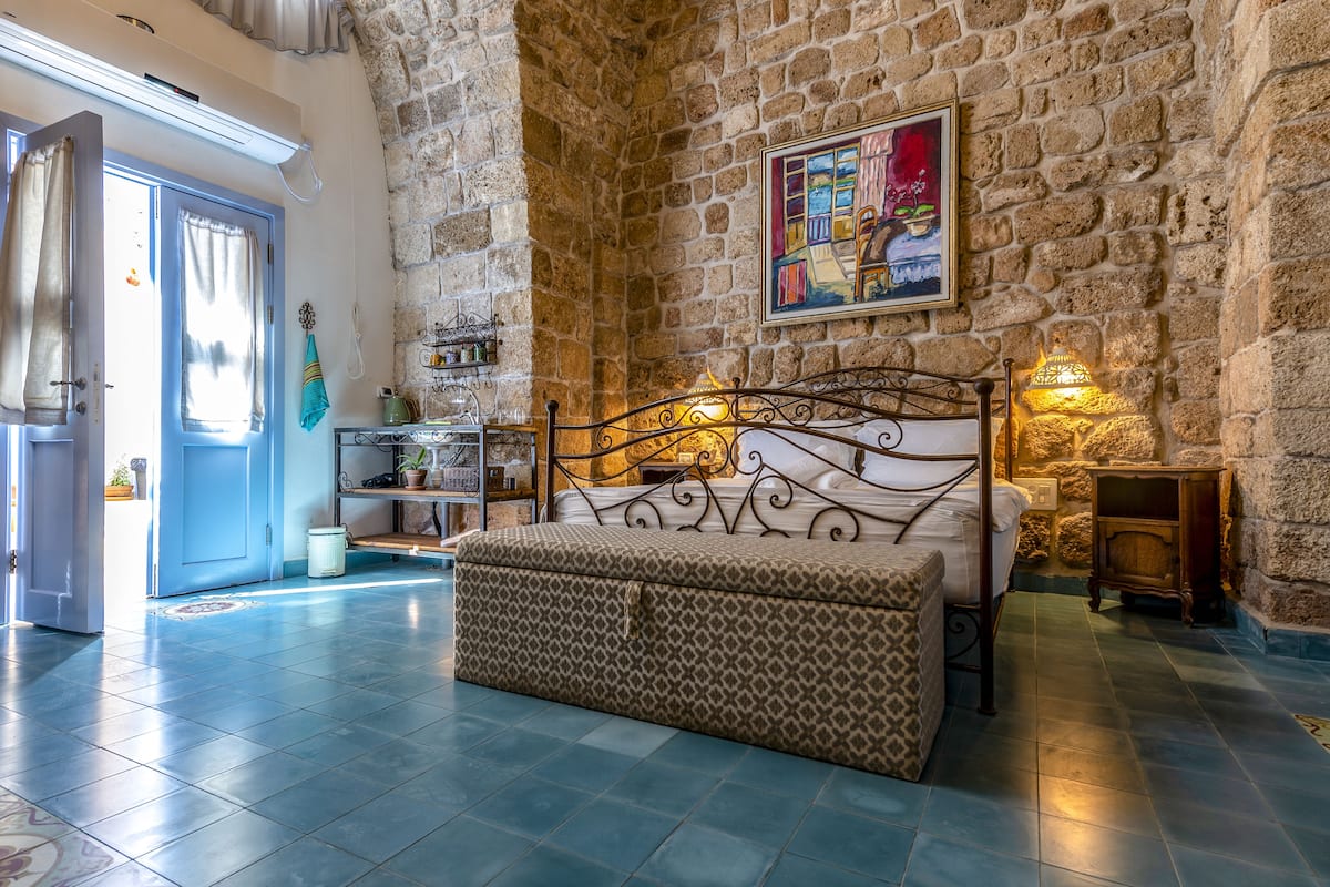 A bedroom with a stone wall, a bed with a patterned headboard, a bedside table with a lamp, and a window with curtains.