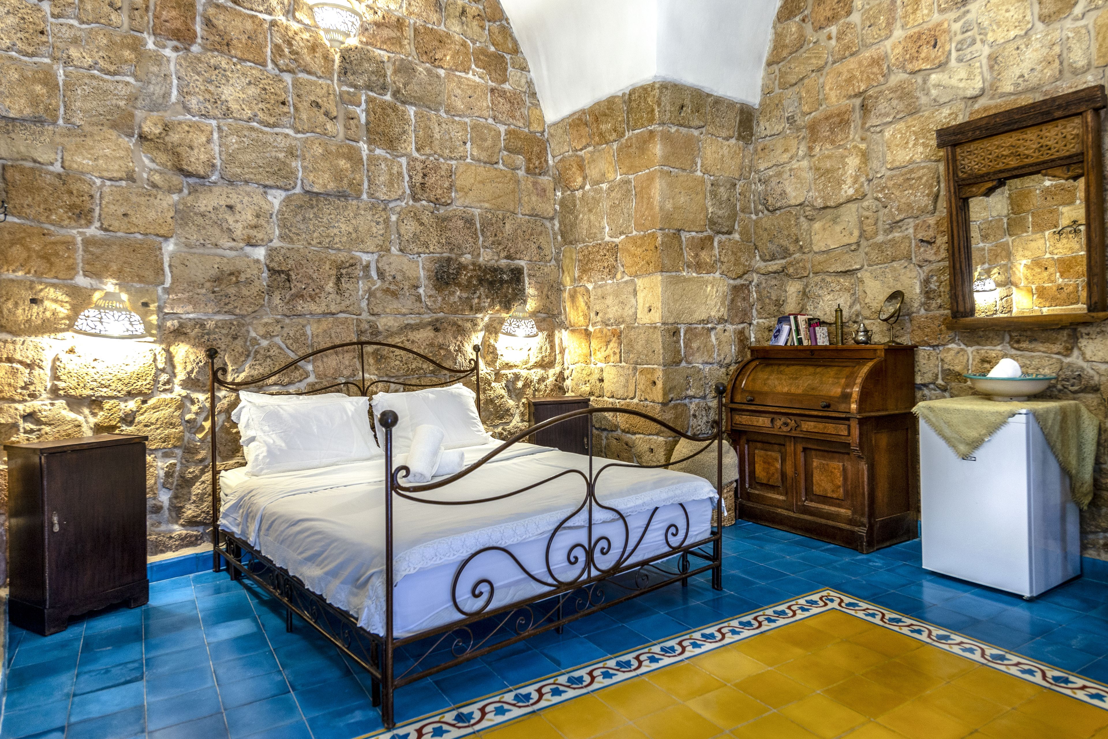 A stone-walled bedroom with a metal bed frame, a wooden nightstand, a white refrigerator, and a patterned rug.