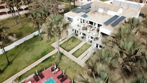 An aerial view of a modern building with a rooftop solar panel array, a swimming pool, and surrounding palm trees.