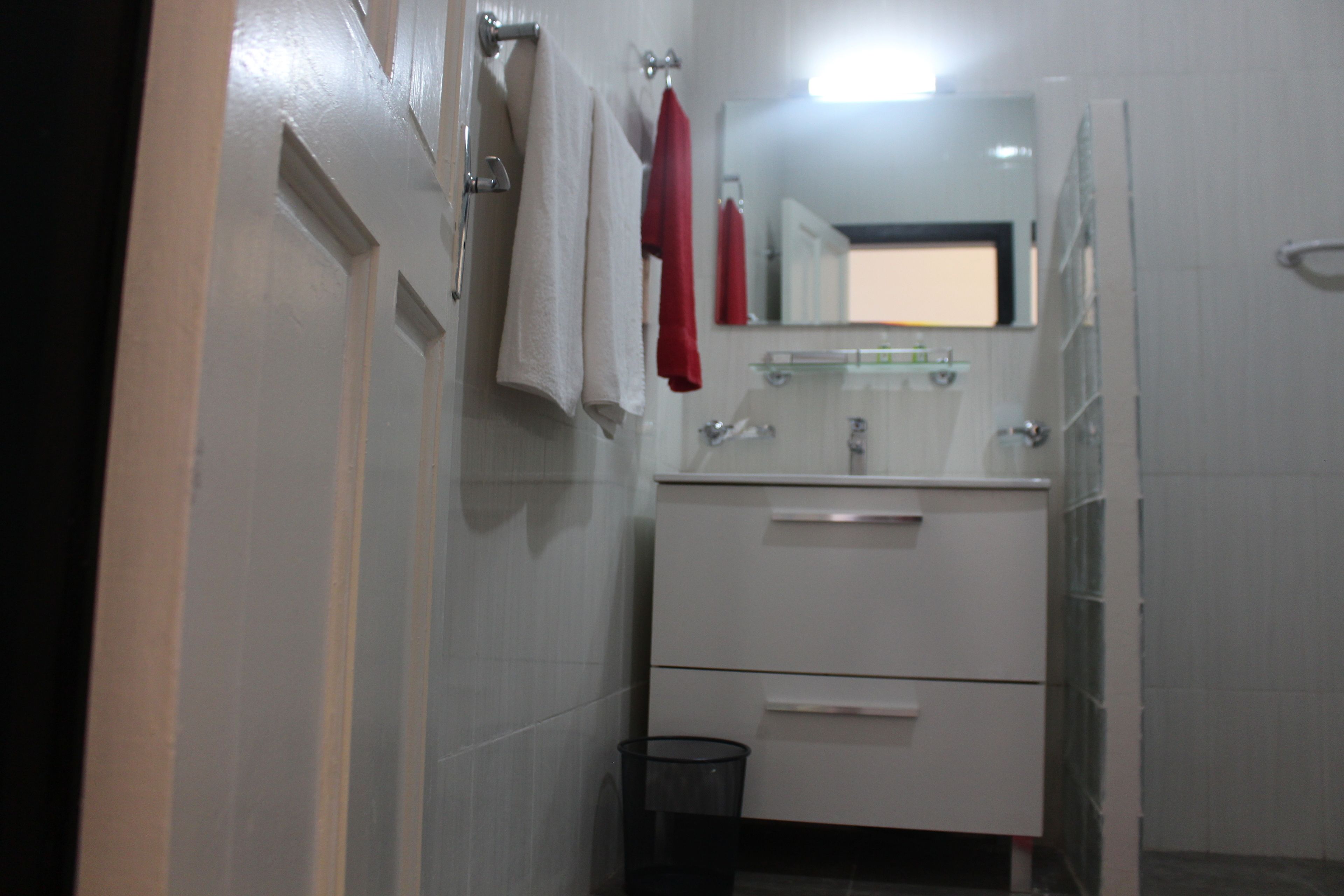 Un baño con paredes de azulejos blancos, un lavabo blanco con un espejo encima y un mueble blanco debajo. Hay dos toallas colgadas en un perchero, una roja y una blanca.