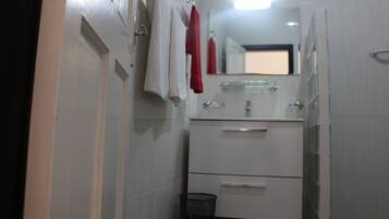 Un baño con paredes de azulejos blancos, un lavabo blanco con un espejo encima y un mueble blanco debajo. Hay dos toallas colgadas en un perchero, una roja y una blanca.