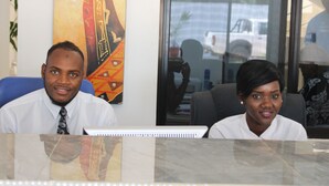 Two individuals seated at a table with a laptop and a notepad, in an office setting with a large window and a colorful painting on the wall.