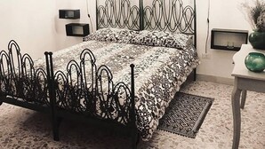 A bedroom with a metal bed frame, patterned bedding, a small bedside table, a nightstand, and a wall-mounted shelf. - B&B Miseno23 (Bacoli)