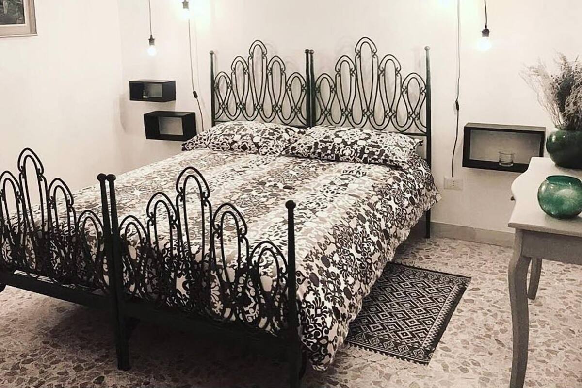 A bedroom with a metal bed frame, patterned bedding, a small bedside table, a nightstand, and a wall-mounted shelf.