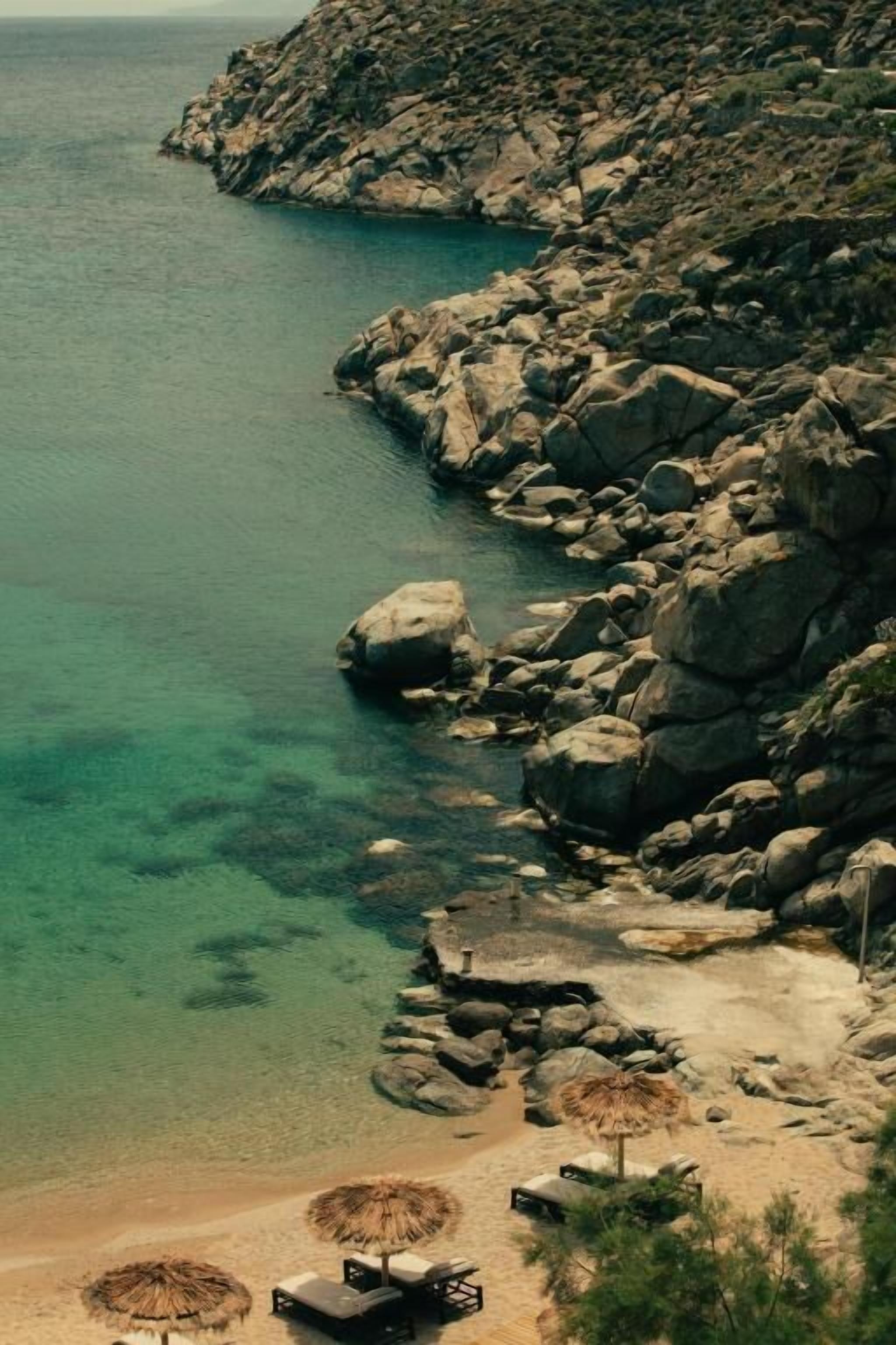 Paisaje costero con agua cristalina, terreno rocoso y una playa con sillas y sombrillas de playa.