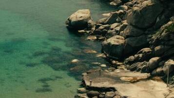 Paisaje costero con agua cristalina, terreno rocoso y una playa con sillas y sombrillas de playa.