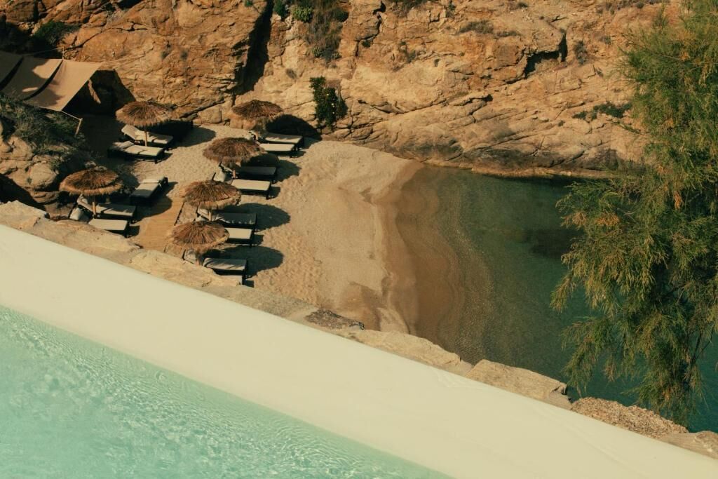 Eine Luftaufnahme eines Strandresorts mit Liegestühlen, Sonnenschirmen, einem Pool und einer felsigen Klippe im Hintergrund.