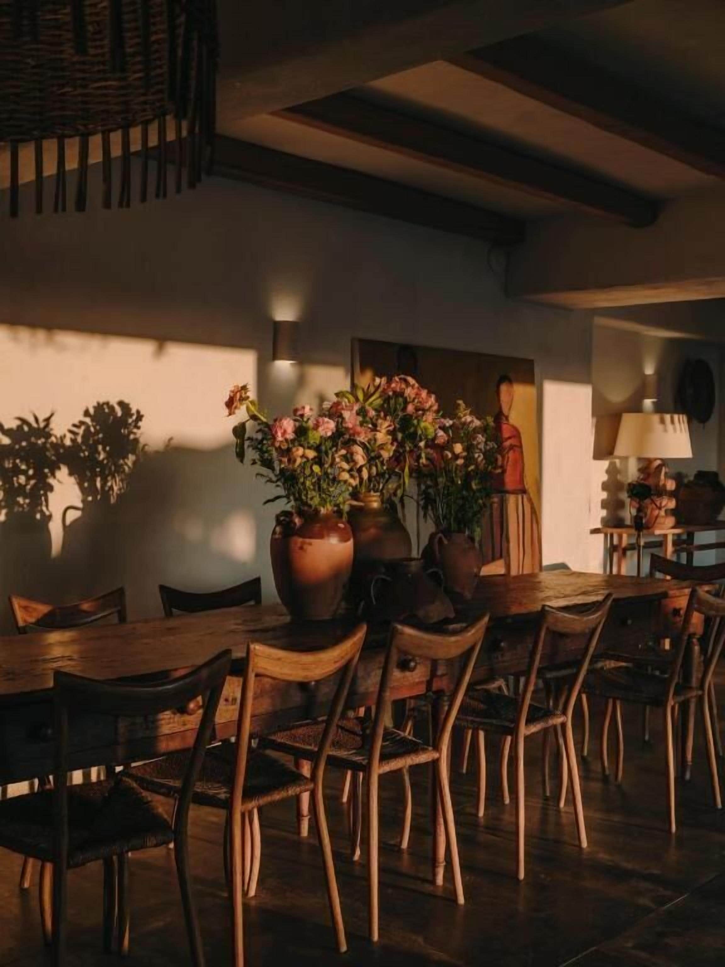 A dining area with a wooden table and chairs, surrounded by potted plants and a vase of flowers.