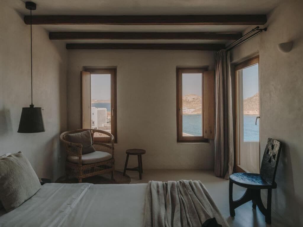 A bedroom with a bed, two windows, a chair, a small stool, and a hanging light fixture.