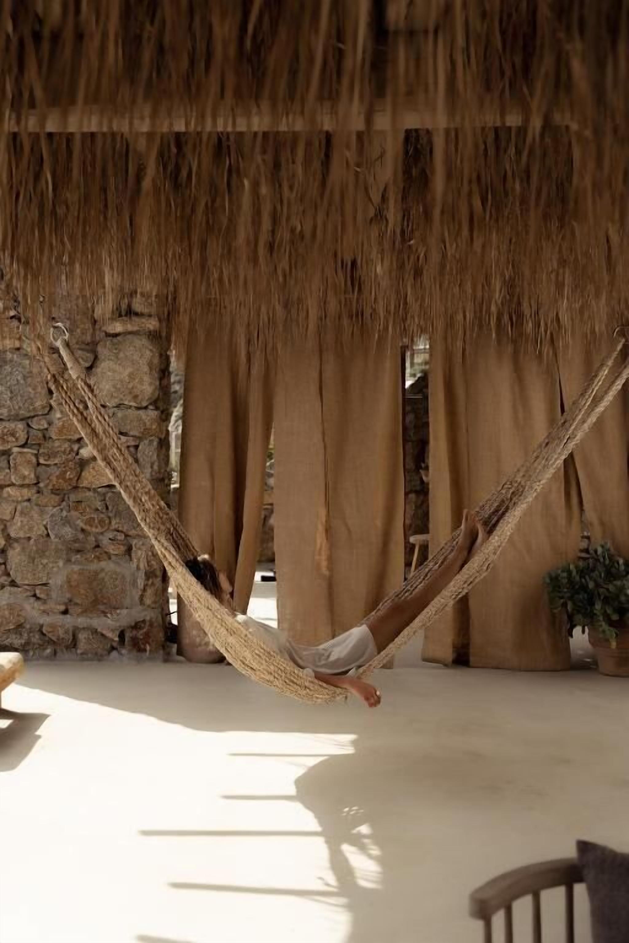 A hammock under a thatched roof, a stone wall, and beige curtains.