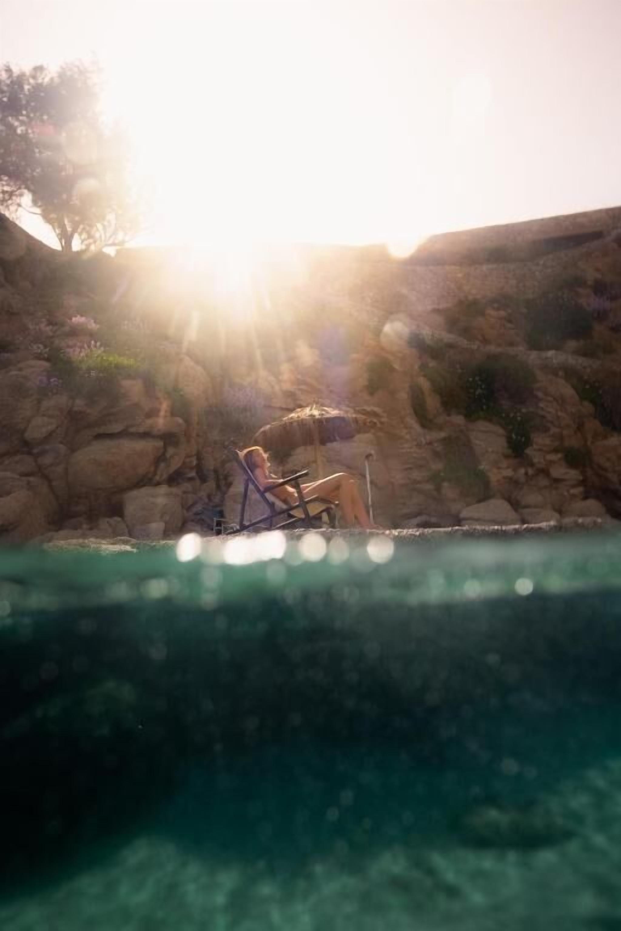 Une personne assise sur une chaise près d’une piscine, avec un paysage rocheux en arrière-plan et la lumière du soleil.